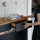 A woman in a black dress opens a black drawer in a modern cupboard with a wooden top. A stack of books and a silver lamp rest on the cupboard surface against a light-coloured wall.