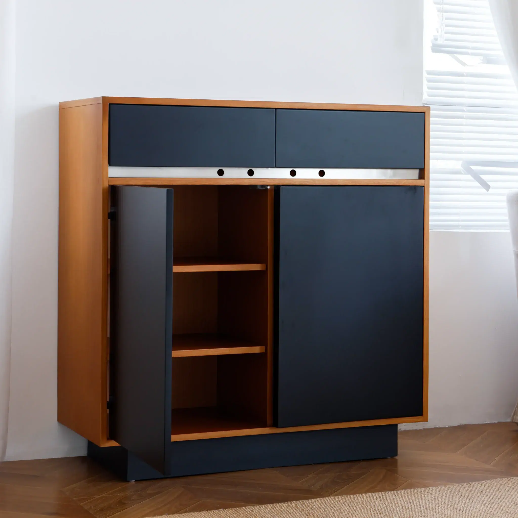 A modern wooden cupboard with black doors and drawers, one door open to reveal two shelves inside, standing on a wooden floor near a white wall and window with blinds.