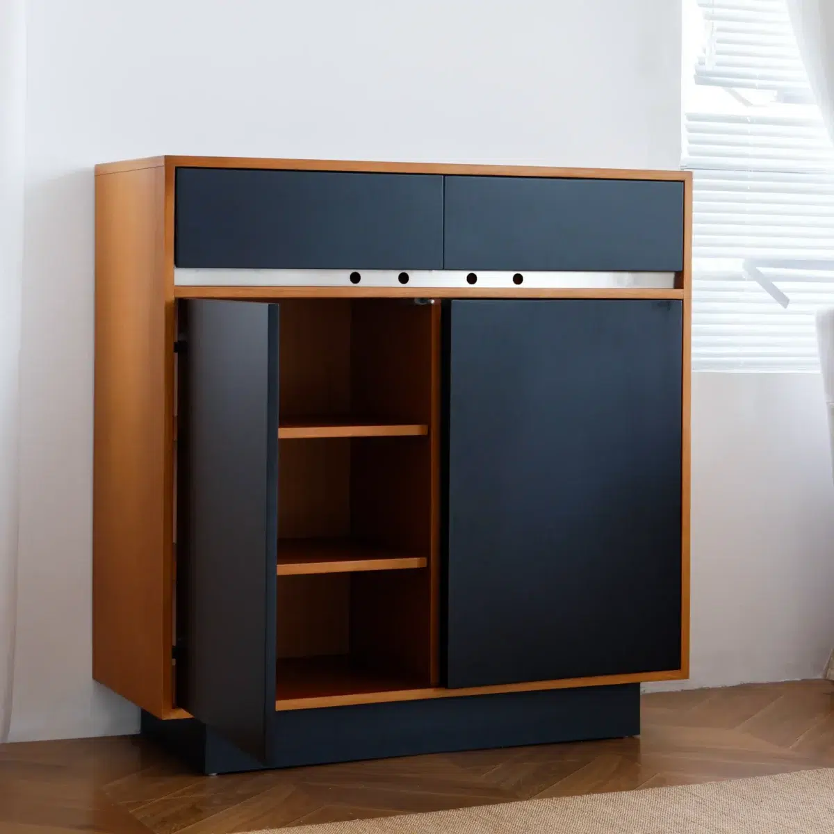 Elly Cabinet 2.0 A modern wooden cupboard with black doors and drawers, one door open to reveal two shelves inside, standing on a wooden floor near a white wall and window with blinds.