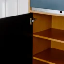An open wooden cupboard with a black door, revealing three empty shelves inside. The cupboard has metal hinges and is set against a white wall.