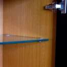 Close-up view of the inside of a wooden cupboard, showing a glass shelf supported by metal pegs and a metal hinge attached to the open cupboard door.