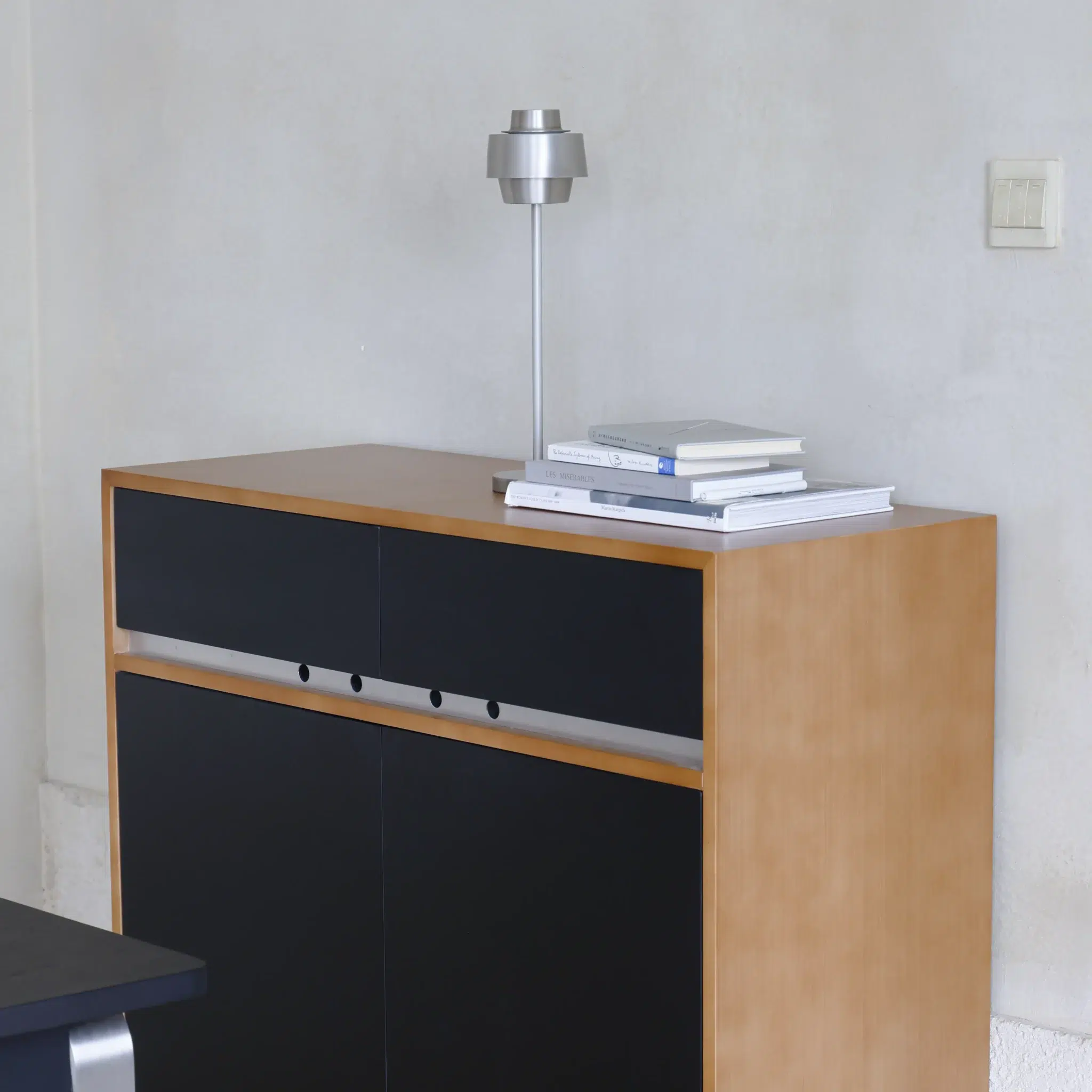 A modern wooden cupboard with black drawer fronts stands against a plain wall, topped with a small silver lamp and several neatly stacked books. An electrical switch is visible on the wall nearby.