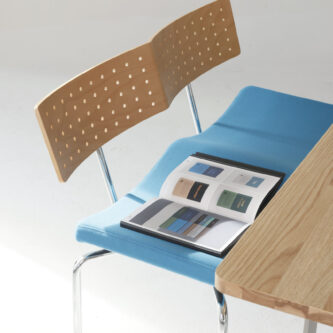 A modern bench with perforated timber backrest, blue fabric seat and slim metal legs and frame, shown beside a wooden table with an open magazine placed on seat.