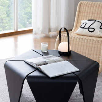 Black sculptural coffee table styled with an open book, candle and lantern light, placed in a modern living room with a wicker lounge chair and large windows.
