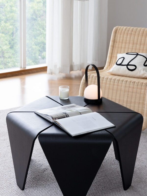 Black sculptural coffee table styled with an open book, candle and lantern light, placed in a modern living room with a wicker lounge chair and large windows.