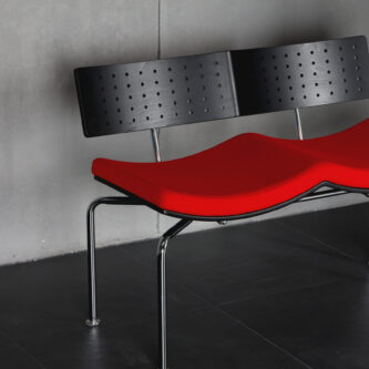 Modern bench seat with perforated black backrest, red fabric seat and metal legs, shown against a concrete wall.