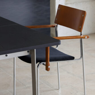 Dining chair with tan timber backrest and armrests, dark fabric seat and steel legs and frame sitting at a black dining table with a light minimal tiled background.
