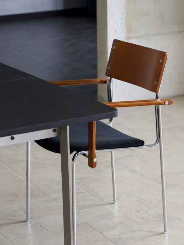 Dining chair with tan timber backrest and armrests, dark fabric seat and steel legs and frame sitting at a black dining table with a light minimal tiled background.
