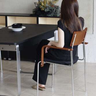 Woman sitting on a timber backrest, steel frame and dark fabric seat dining chair with her back to camera, beside a dark wooden table and cabinet with flowers, on a light tiled floor.