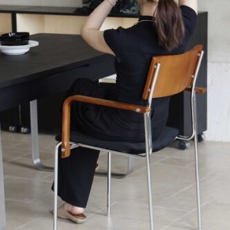 Woman sitting on a timber backrest, steel frame and dark fabric seat dining chair with her back to camera, beside a dark wooden table on a light tiled floor.
