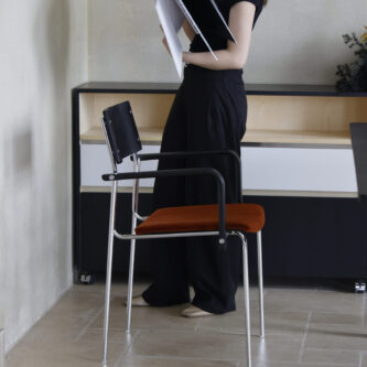 Woman standing beside a dining chair with a timber back, steel frame and tan fabric seat, holding an open book in a minimalist room.