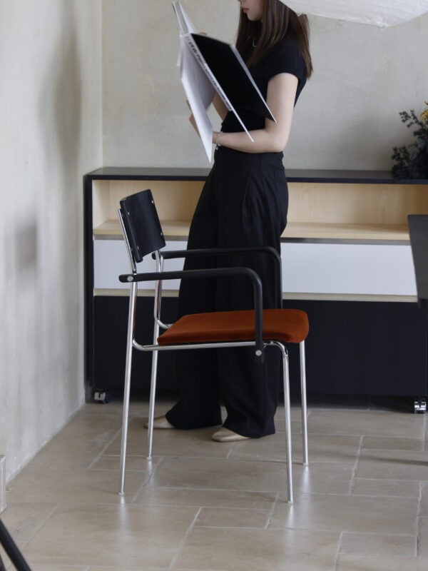 Woman standing beside a dining chair with a timber back, steel frame and tan fabric seat, holding an open book in a minimalist room.