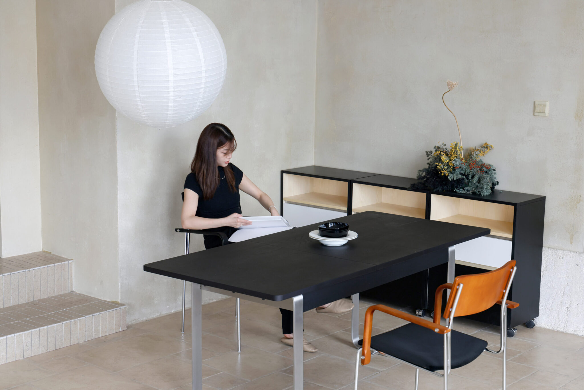 A woman sits at a black table in a minimalist room, reading a book. There is a paper lantern overhead, a shelf with a plant arrangement behind her, and two chairs at the table. The walls and floor are light-coloured.