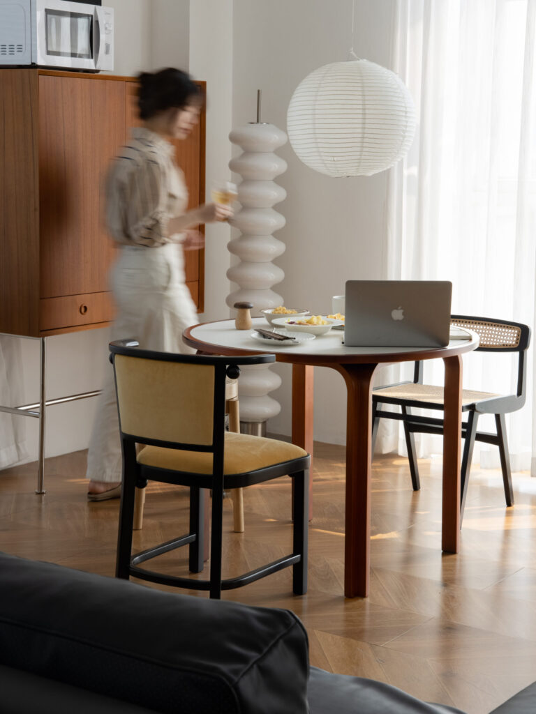 A blurred woman stands near a round dining table with a laptop, plates of food, and a cup. The room has wooden furniture, a paper lantern, and natural light streaming through sheer curtains.