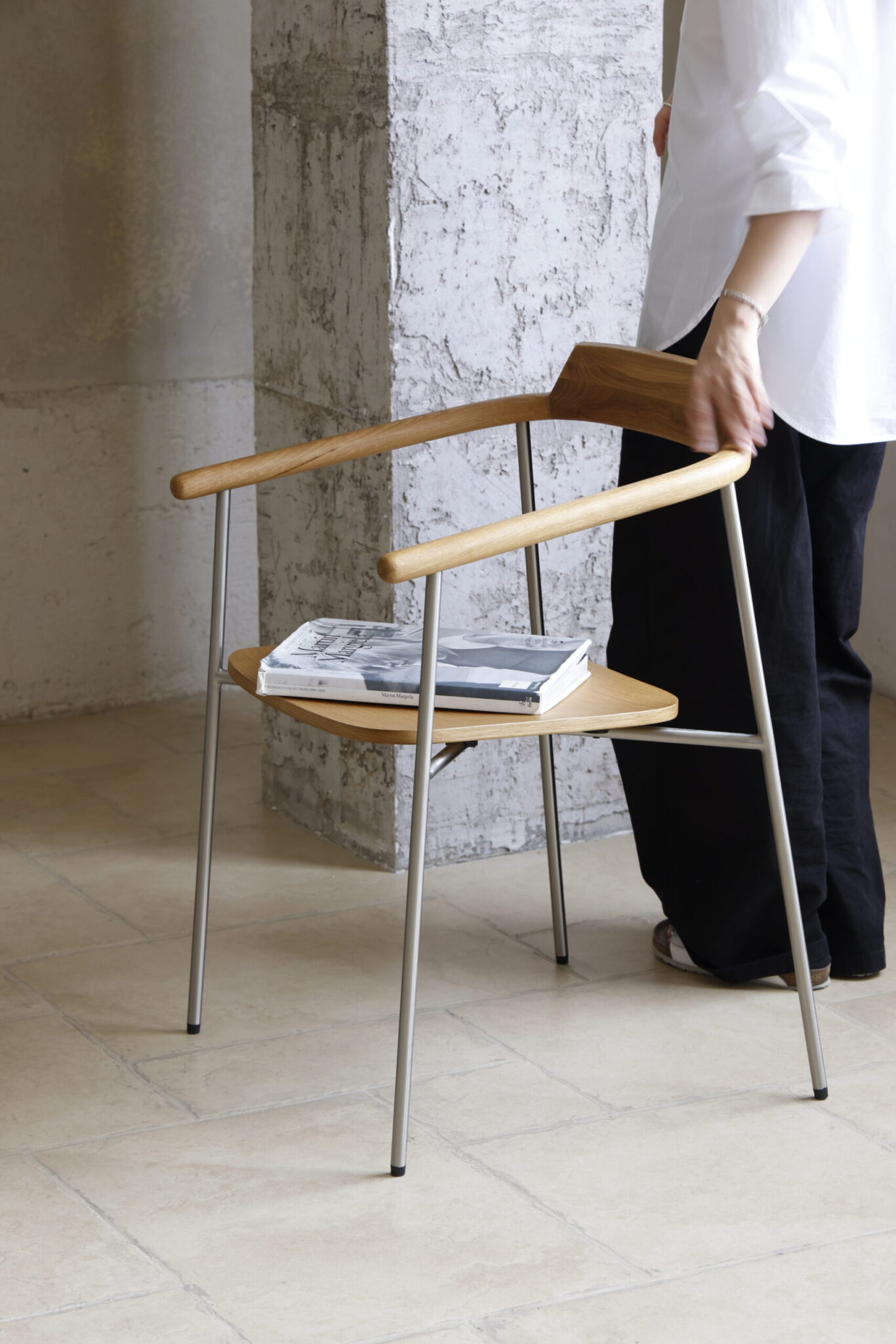 A person in a white shirt and black trousers stands next to a modern wooden chair with metal legs, which has a folded newspaper placed on the seat. The scene is indoors with neutral-toned tiled flooring.