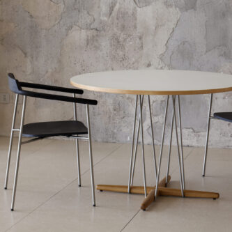 Modern round table with a light laminate top, slender metal legs and a timber cross-base, paired with black dining chairs in a textured interior.