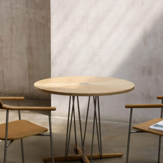 Modern round timber table with slender metal legs and a cross-base, shown in a styled interior with matching timber chairs.