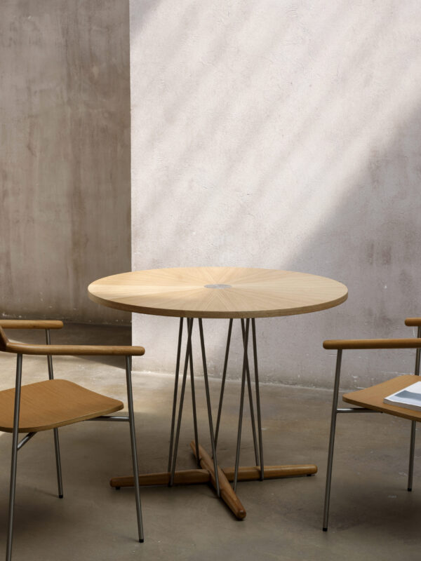 Modern round timber table with slender metal legs and a cross-base, shown in a styled interior with matching timber chairs.