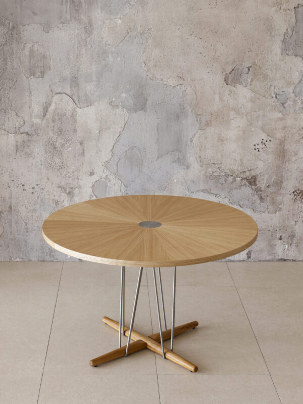 Modern round timber table with slender metal legs and a cross-base, shown in front of a concrete wall.