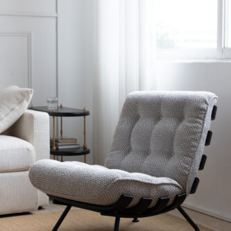 Light grey tufted lounge chair with black metal strap frame, styled in a bright white living room beside a cream sofa and a small side table, with soft natural light coming through sheer curtains.