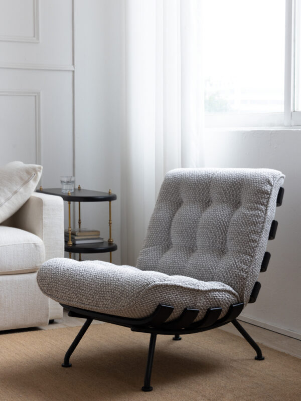 Light grey tufted lounge chair with black metal strap frame, styled in a bright white living room beside a cream sofa and a small side table, with soft natural light coming through sheer curtains.