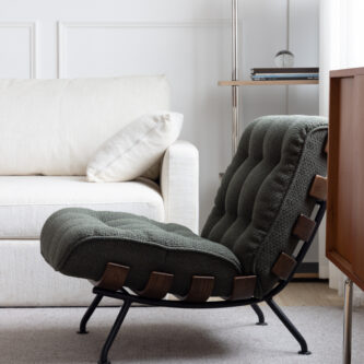 Side-view of a green tufted lounge chair with wooden strap detailing, styled in a light, modern living room beside a cream sofa, metal-framed shelf, and soft natural window light.