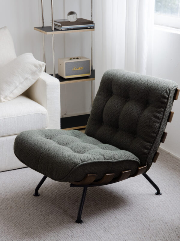 Green tufted lounge chair with wooden strap detailing, styled in a light, modern living room beside a cream sofa, metal-framed shelf, and soft natural window light.