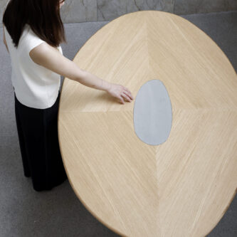 Overhead view of rounded wooden dining table with metal cut-out in centre, shown on minimal concrete background with women caressing centre.