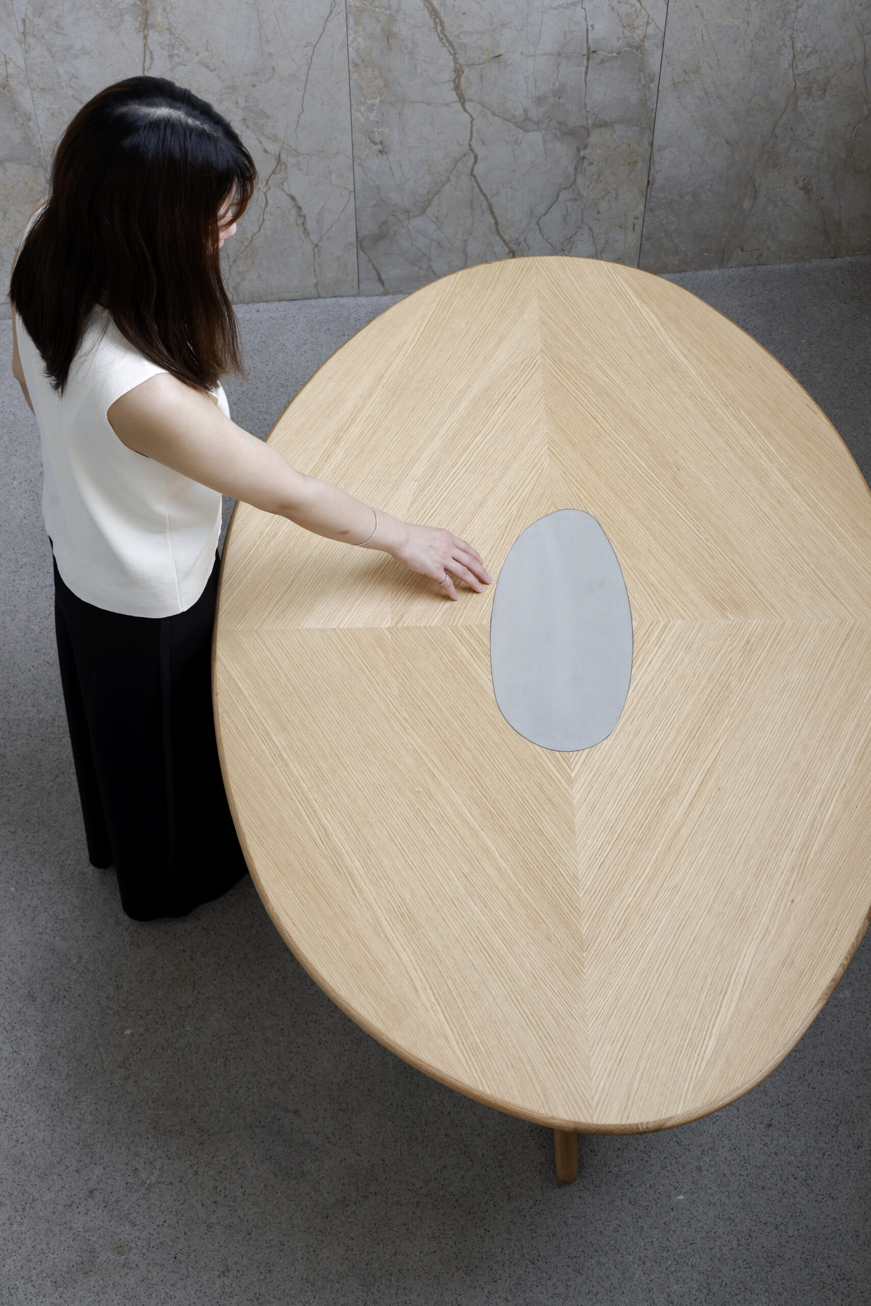 Overhead view of rounded wooden dining table with metal cut-out in centre, shown on minimal concrete background with women caressing centre.