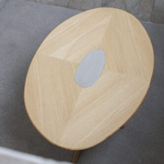 Overhead view of rounded wooden dining table with metal cut-out in centre, shown on minimal concrete background.
