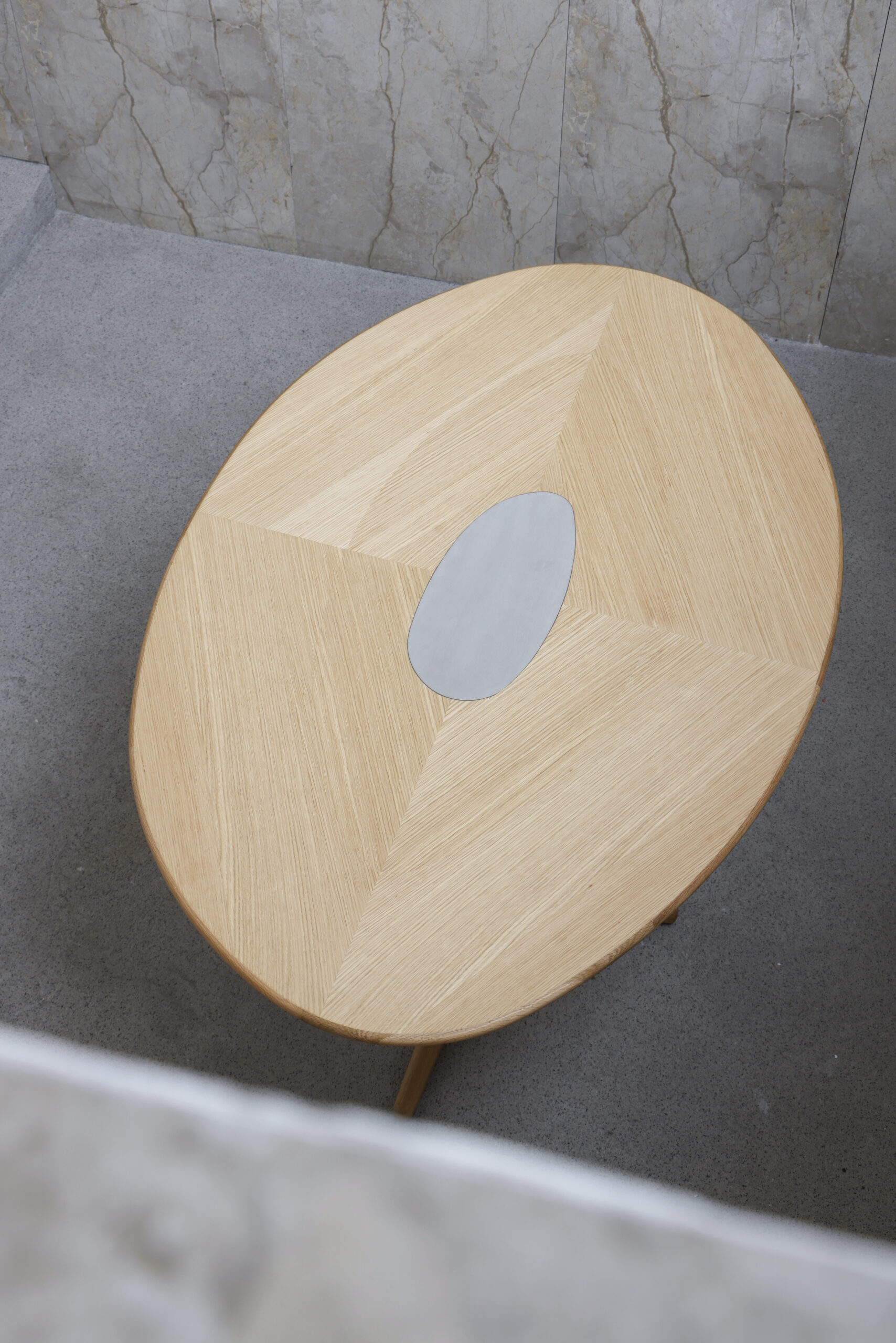Pebble Dining Table Overhead view of rounded wooden dining table with metal cut-out in centre, shown on minimal concrete background.