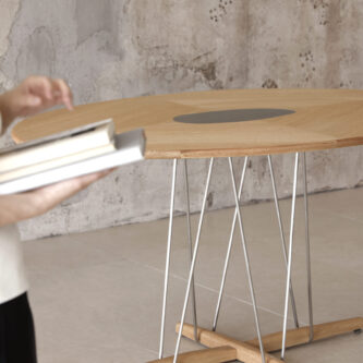 Close-up image of rounded wooden dining table, sculptural metal legs and timber top against concrete wall, person holding books in forefront.