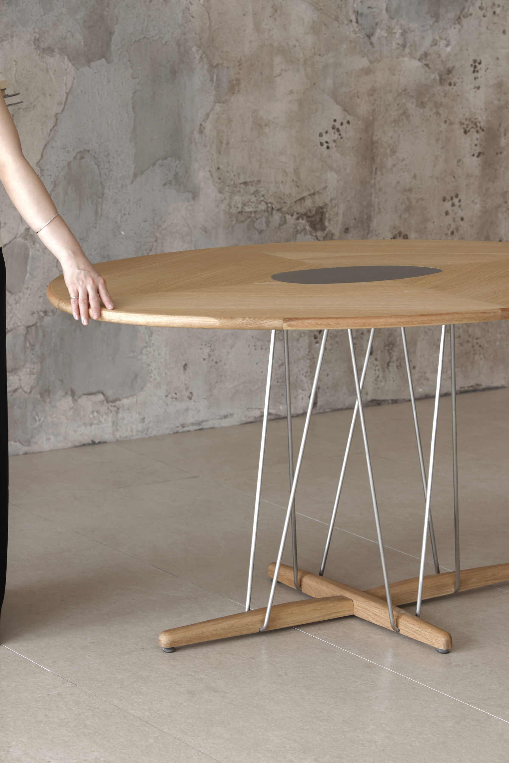 Pebble Dining Table Close-up image of rounded wooden dining table, sculptural metal legs, and hand resting on timber top against concrete wall.