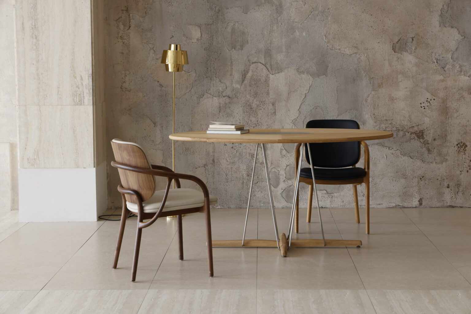 A modern, minimalist room with a round wooden table, two chairs—one with a black seat and back and one with a light cushion—stacked books, a gold floor lamp, and a textured, weathered wall.