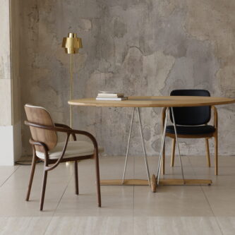 Minimalist dining setup featuring a round wooden table with stainless steel crossed legs, a brass lamp and two wooden chairs against a concrete wall.