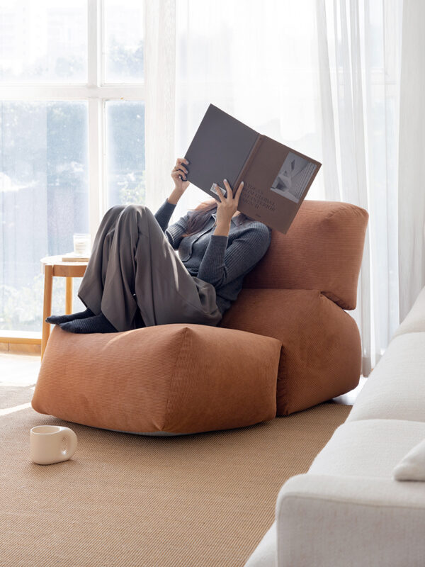 A woman sits on a sculptural brown cushioned lounge chair with an integrated extended seat, reading a book which hides her face, shown in a light living room.