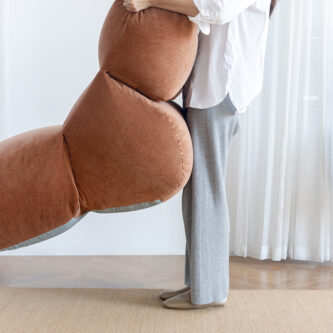 A woman lifts a sculptural brown cushioned lounge chair, showing its lightweight design, shown in a minimal living room.