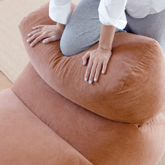 A woman presses her hands onto a sculptural brown lounge chair with an integrated extended seat to show its cushioned structure.