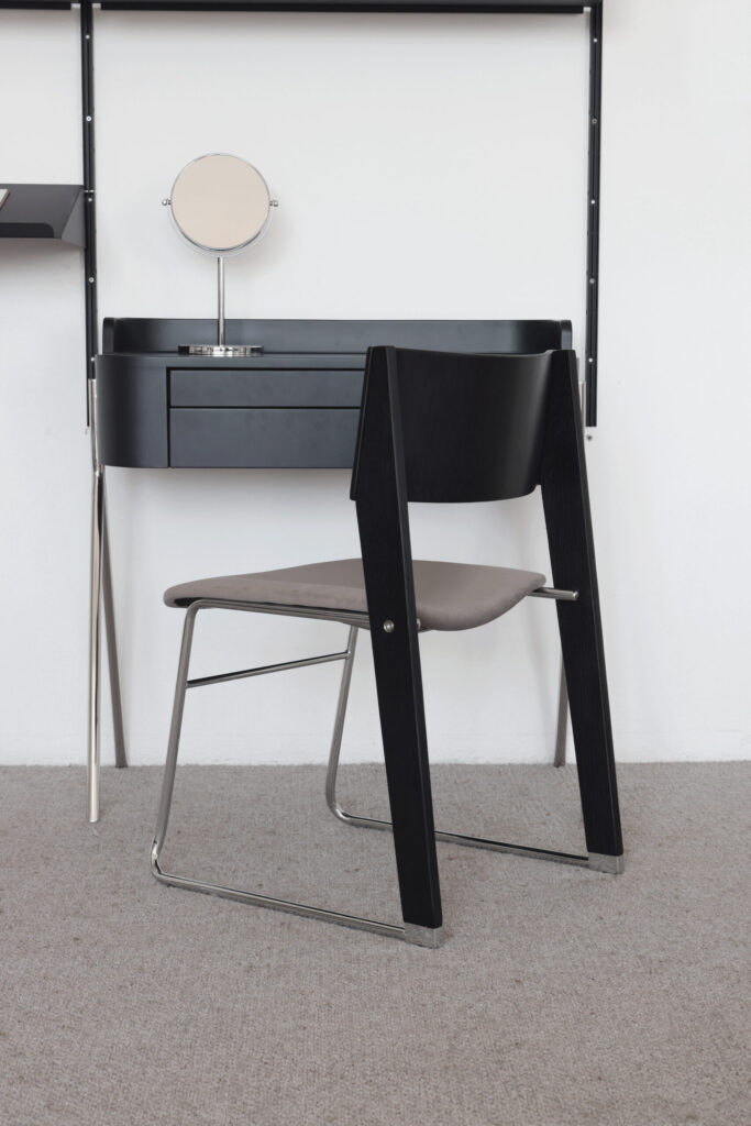 A modern black dressing table with a round mirror and drawers, paired with a minimalist chair featuring black wooden sides and metal legs, set against a white wall and beige carpet.