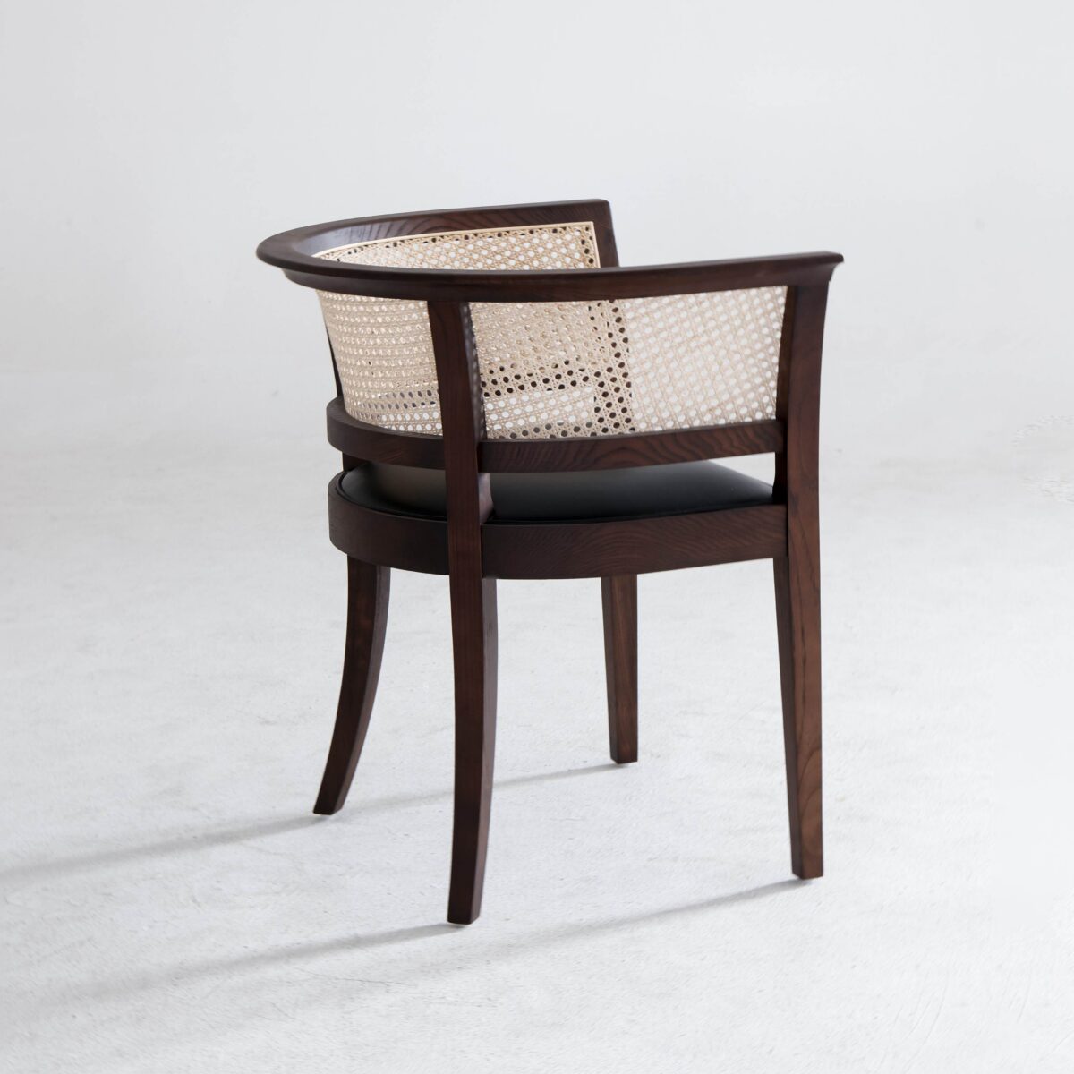 Faaborg Chair_B1 A wooden chair with a curved back and woven cane backrest sits on a light-coloured floor. The seat is black and cushioned, and the overall design has a classic, elegant look.
