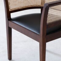 A close-up of a wooden chair with a dark brown frame, a woven cane backrest and side, and a black leather seat, set on a light-coloured floor.