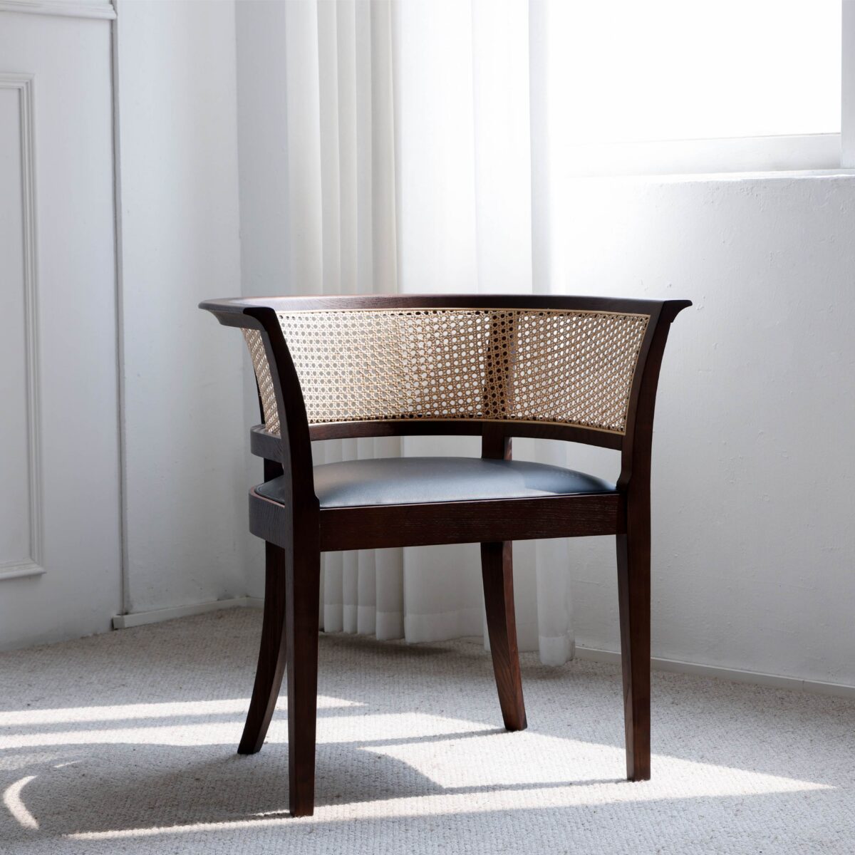 Faaborg Chair_C1 A wooden accent chair with curved cane backrest and dark frame sits in a bright, sunlit room with white walls, carpeted floor, and sheer curtains near a window.