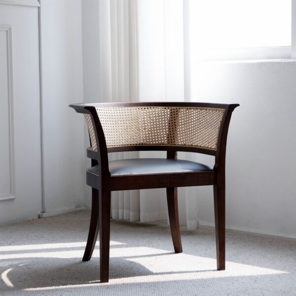A wooden accent chair with curved cane backrest and dark frame sits in a bright, sunlit room with white walls, carpeted floor, and sheer curtains near a window.