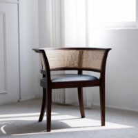 A wooden accent chair with curved cane backrest and dark frame sits in a bright, sunlit room with white walls, carpeted floor, and sheer curtains near a window.