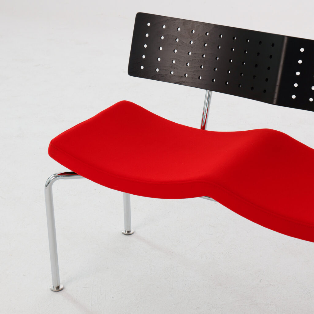 A modern chair with a wavy red seat, a perforated black backrest, and chrome legs, positioned on a white floor.