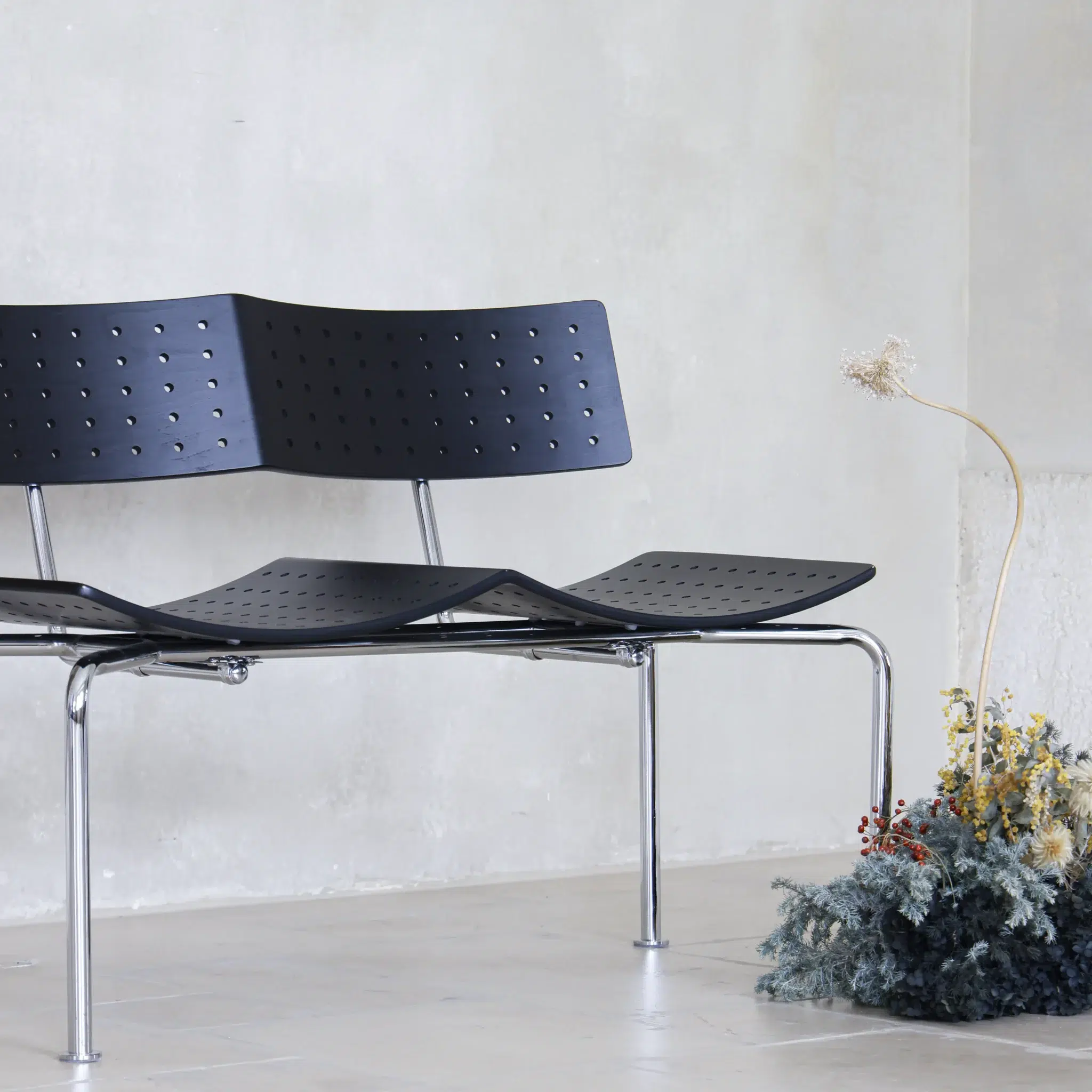 A modern black three-seat bench with perforated seats and chrome legs sits on a light tiled floor beside a minimal arrangement of dried flowers against a plain, light-coloured wall.
