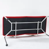 A modern chair with a red edge and metal legs is flipped upside down on a white floor, revealing its black underside and the metal frame supporting the seat.