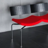 A modern bench with a curved red seat, perforated black backrest, and chrome legs, set against a grey concrete wall and dark tiled floor.