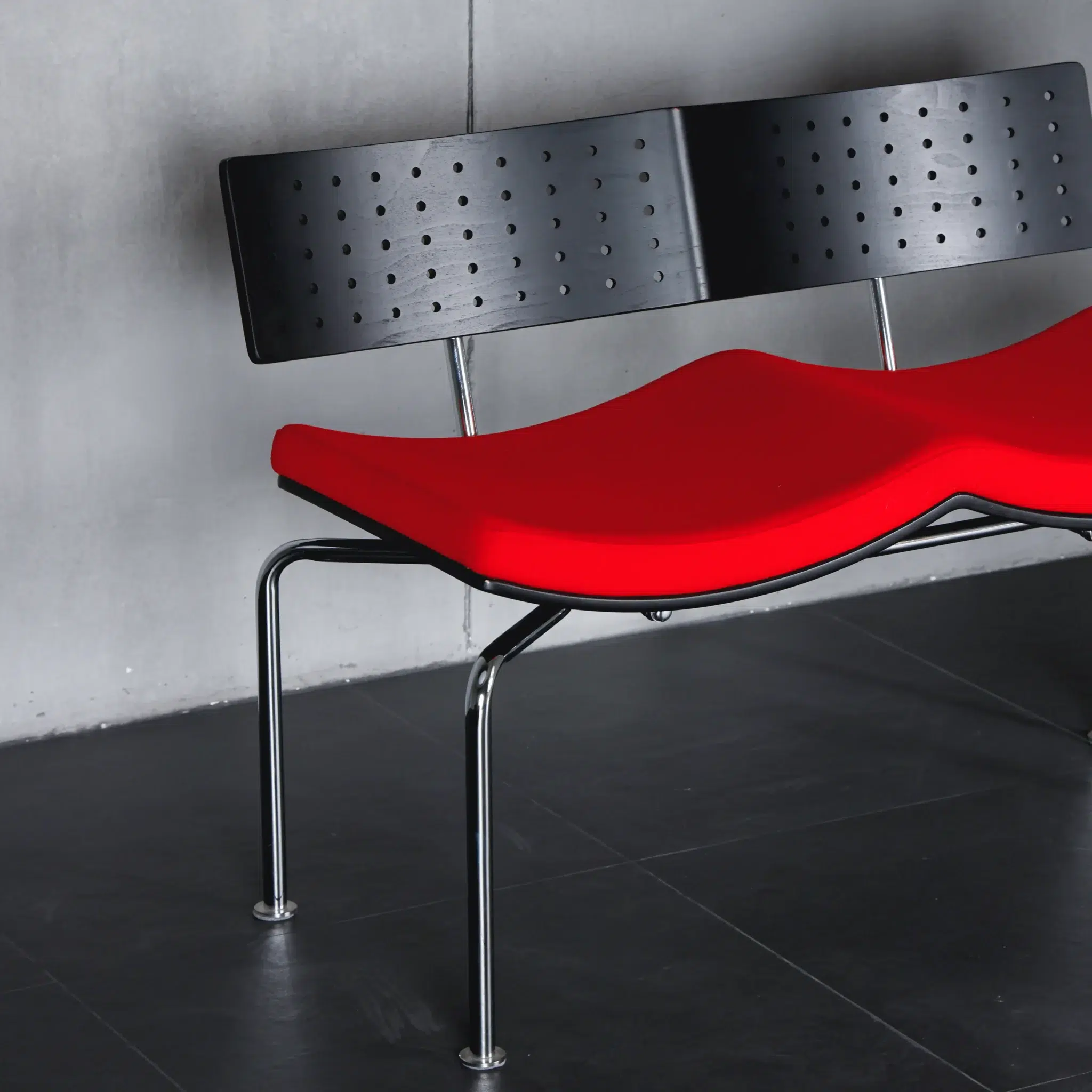 A modern bench with a bright red, contoured seat, black perforated backrest, and metal legs, positioned against a grey wall on a dark tiled floor.