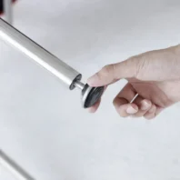 A hand is screwing a round, black-and-silver adjustable foot into the end of a metal tube, likely assembling furniture.